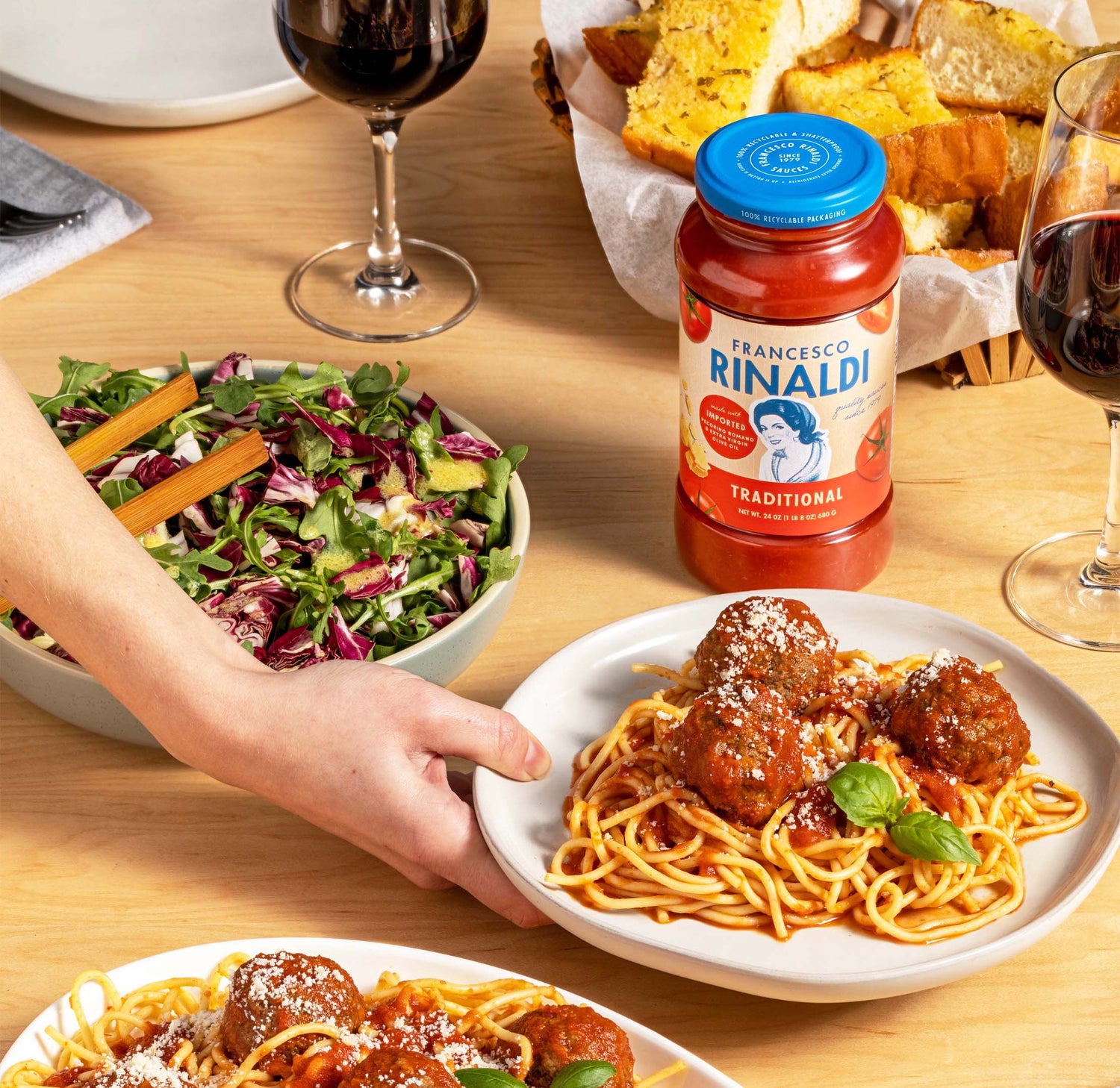 A jar of Francesco Rinaldi sauce next to full plats of salad and spaghetti and two glasses of red wine.